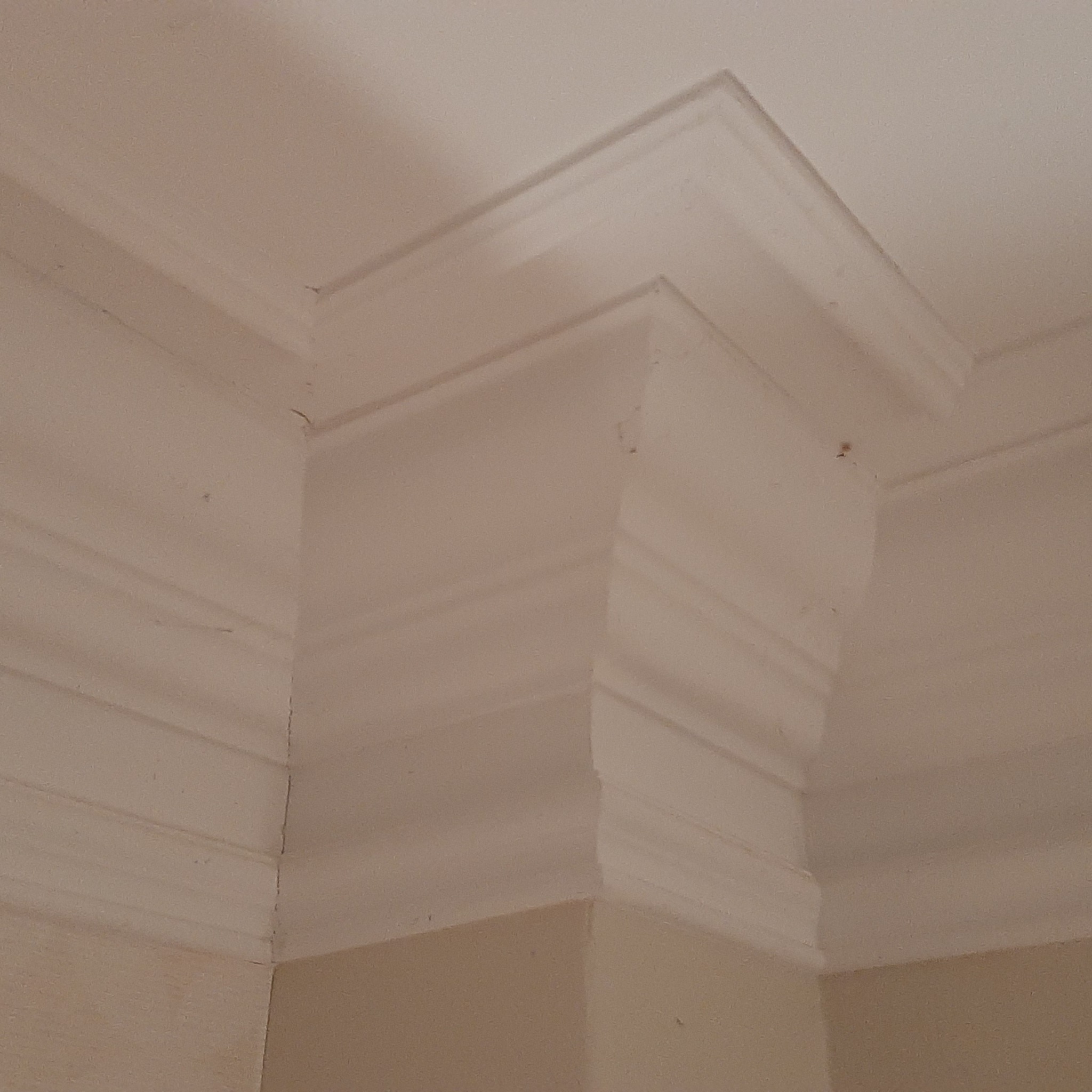 Wykończony narożnik sufitu z listwą dekoracyjną, świeżo pomalowany na biało, widoczne delikatne niedoskonałości na ścianie w kolorze beżowym.