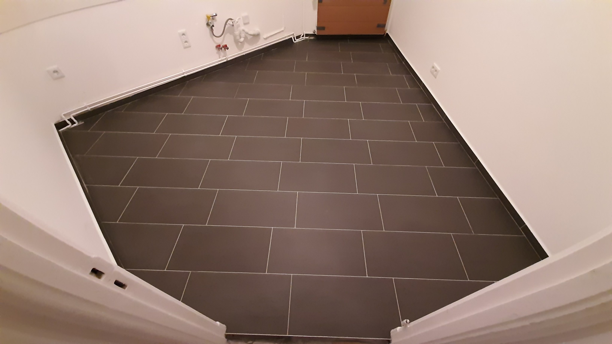 Ukończone układanie ciemnoszarych, prostokątnych płytek podłogowych w pomieszczeniu o białych ścianach, widoczne rury grzewcze przy podłodze, ujęcie z perspektywy wejścia.