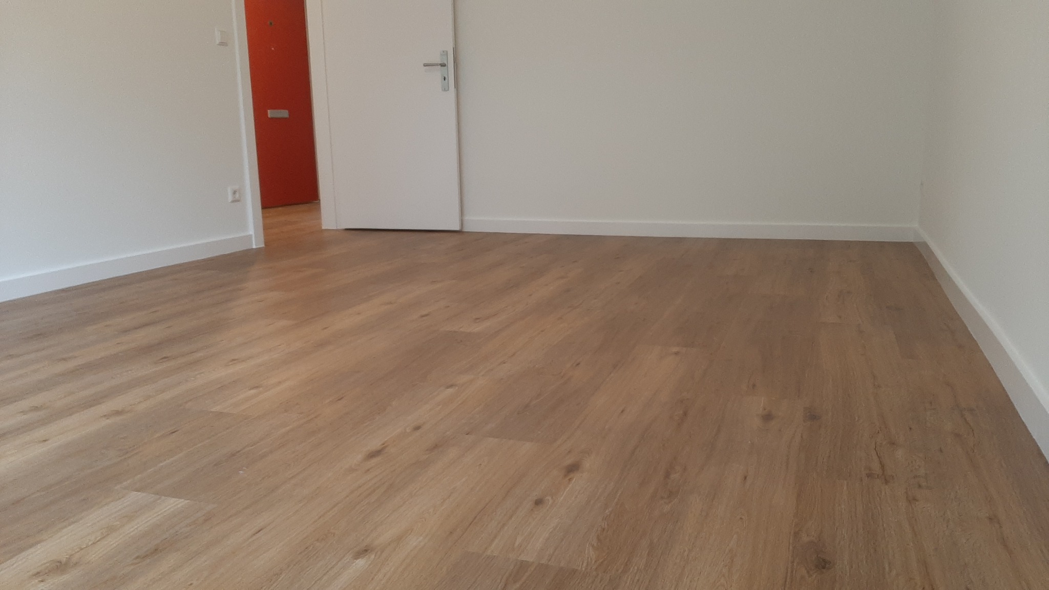 Jasne panele podłogowe imitujące drewno w pustym pokoju z białymi ścianami i pomarańczowymi drzwiami w tle.