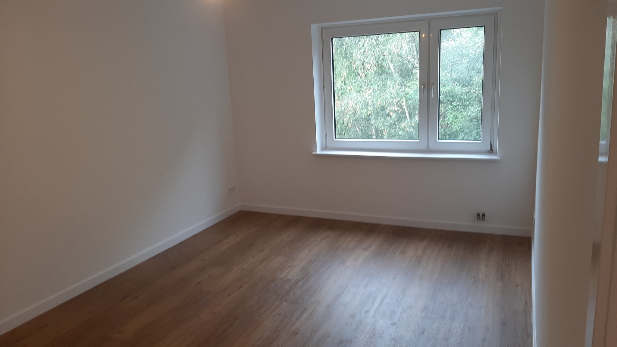 Pusty pokój po remoncie z drewnianą podłogą, białymi ścianami i oknem z widokiem na zieleń, listwy przypodłogowe pomalowane na biało, widoczne gniazdka elektryczne.
