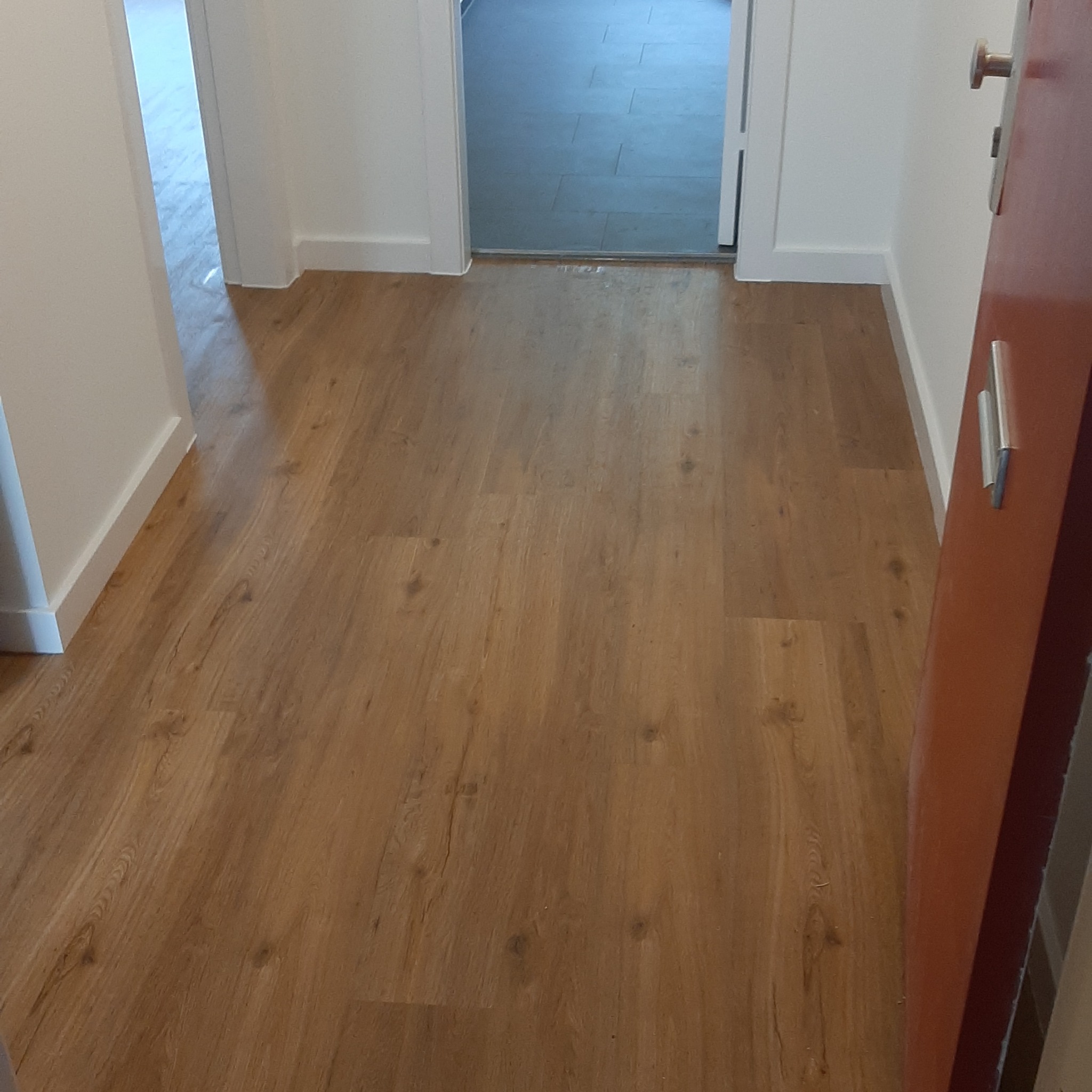 Nowo ułożone panele podłogowe w korytarzu, widok z perspektywy otwartych drzwi, z widocznym fragmentem kolejnego pomieszczenia z szarą podłogą.