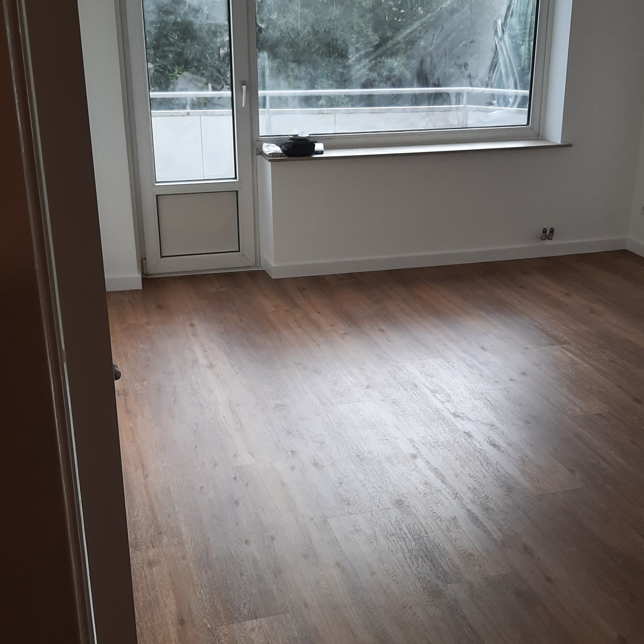 Jasne panele podłogowe z wyraźną strukturą drewna, widok na okno balkonowe i akcesoria na parapecie, fragment ościeżnicy w kadrze.