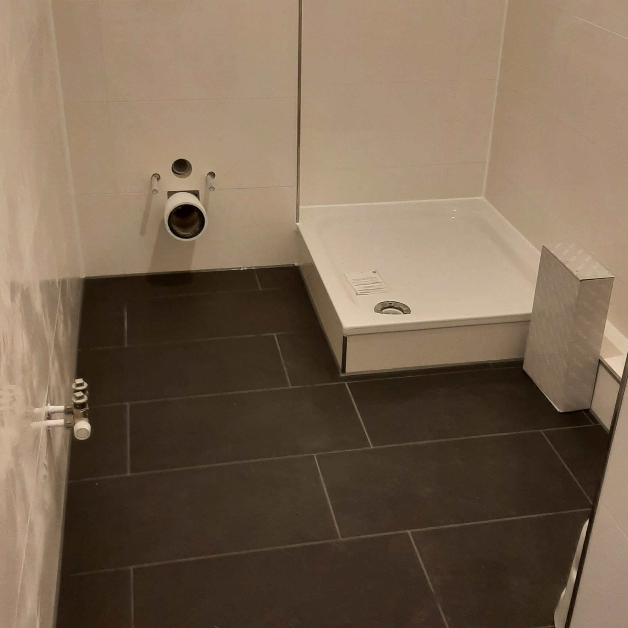 Nowoczesna łazienka w trakcie remontu: ciemne, prostokątne płytki podłogowe, biały brodzik z odpływem, wystające rury i otwór kanalizacyjny w ścianie, kartonowe opakowanie przy brodziku.