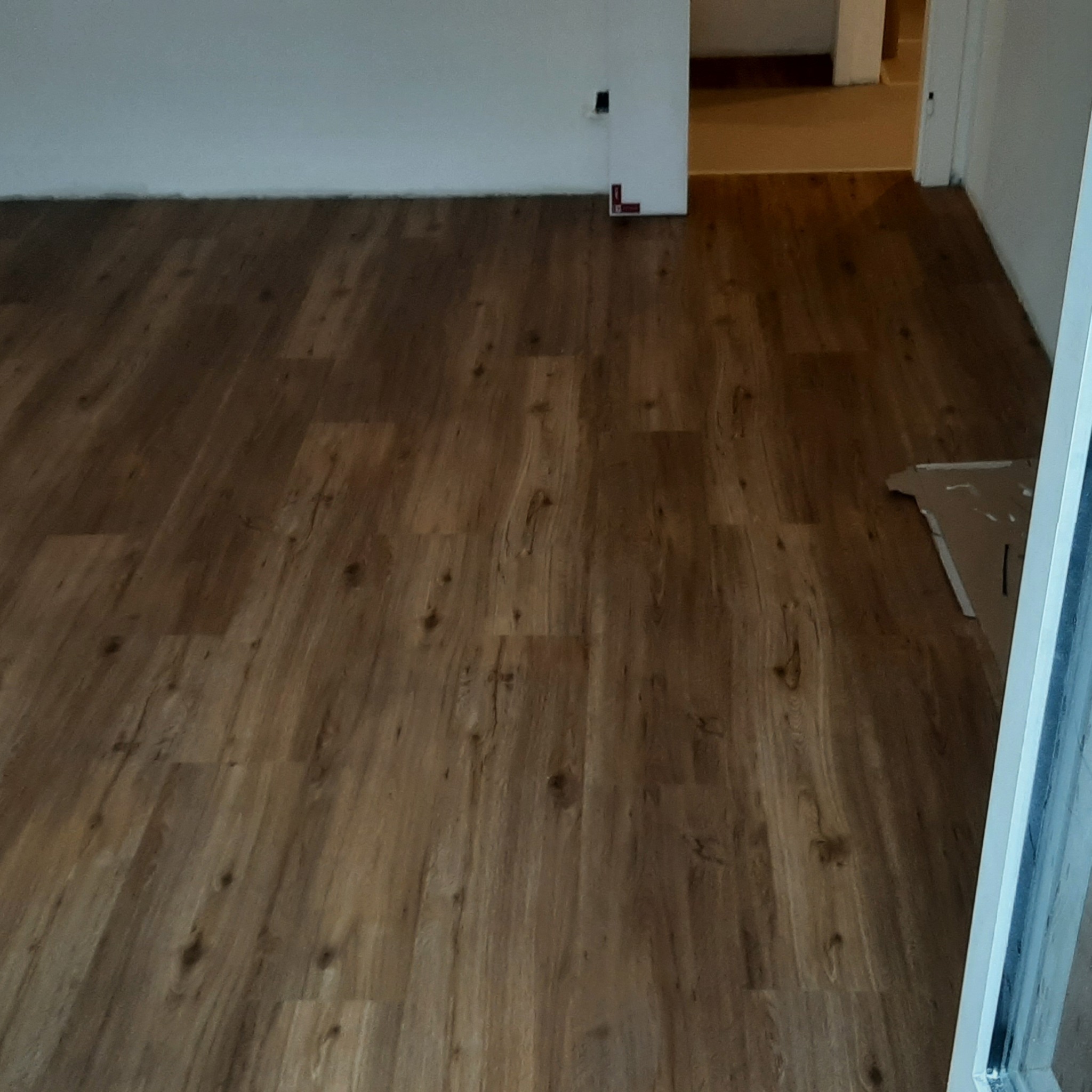 Ułożone panele podłogowe w odcieniach brązu, widoczne łączenia i faktura drewna, fragment kartonu przy ścianie, biała ściana i fragment drzwi w tle.