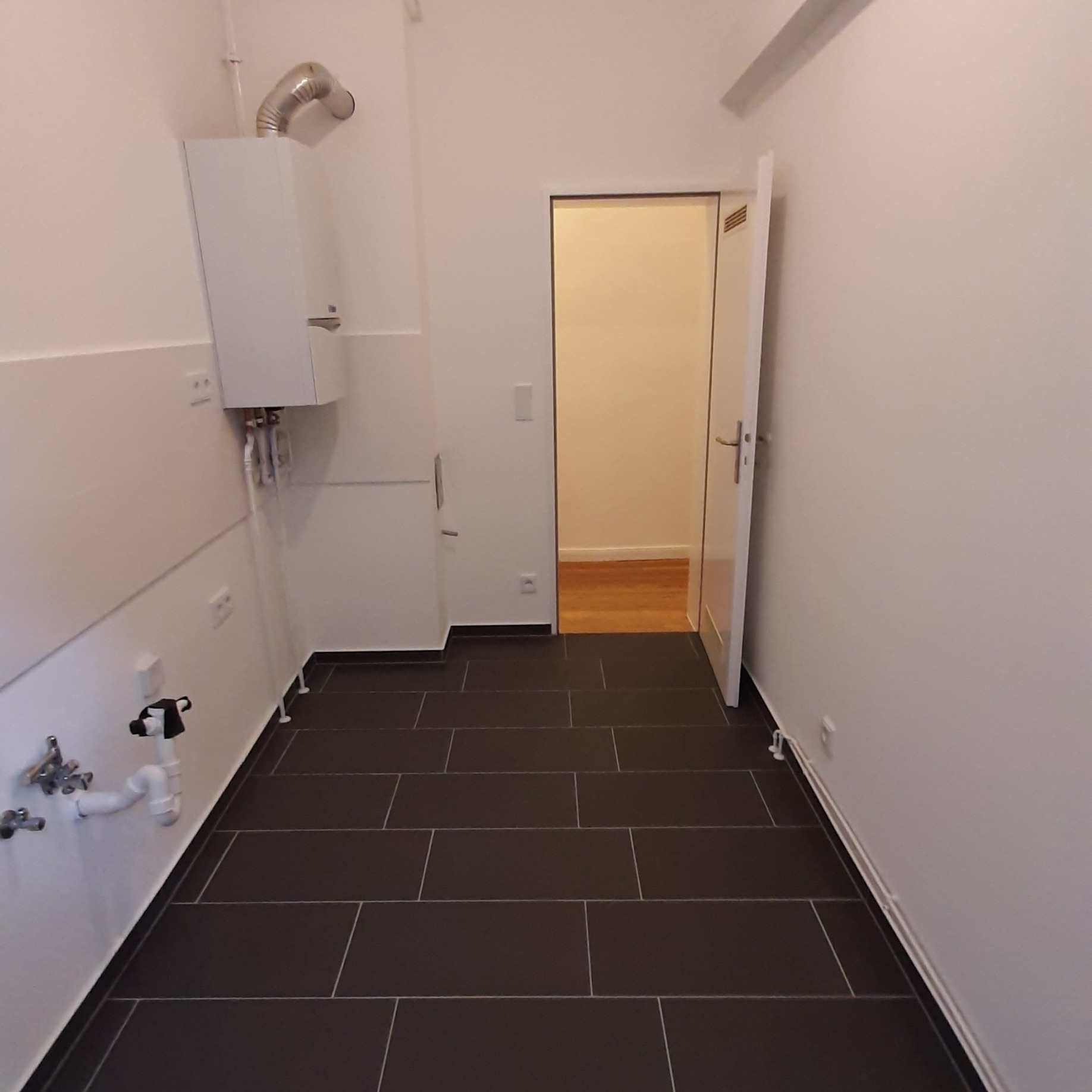 Wnętrze pomieszczenia z ułożoną ciemną, prostokątną terakotą na podłodze, białymi ścianami, grzejnikiem gazowym i otwartymi białymi drzwiami do innego pokoju z drewnianą podłogą.