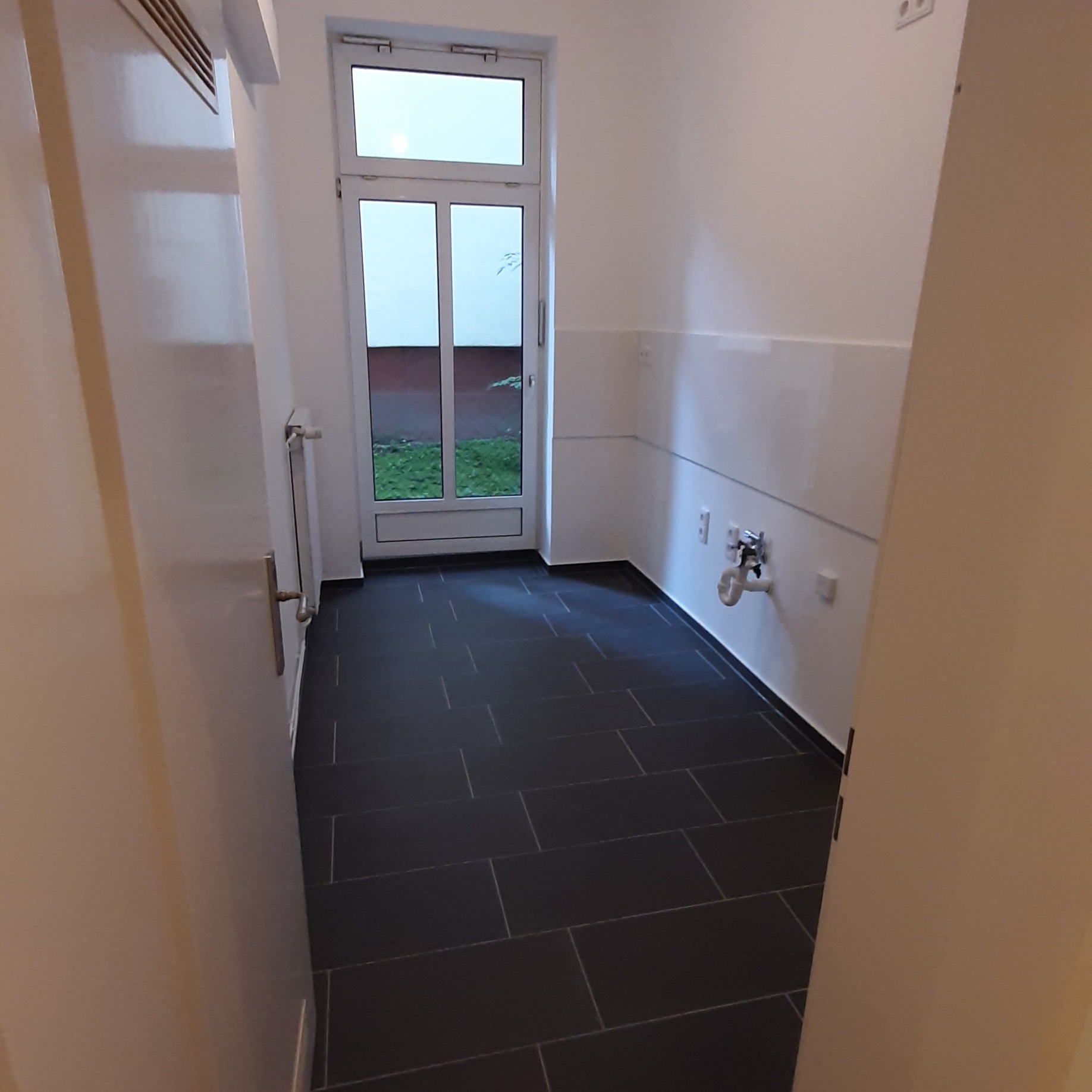 Wykończona łazienka z ciemną, prostokątną glazurą podłogową i białą glazurą ścienną do połowy wysokości, widoczna instalacja wodna i okno z widokiem na zieleń.