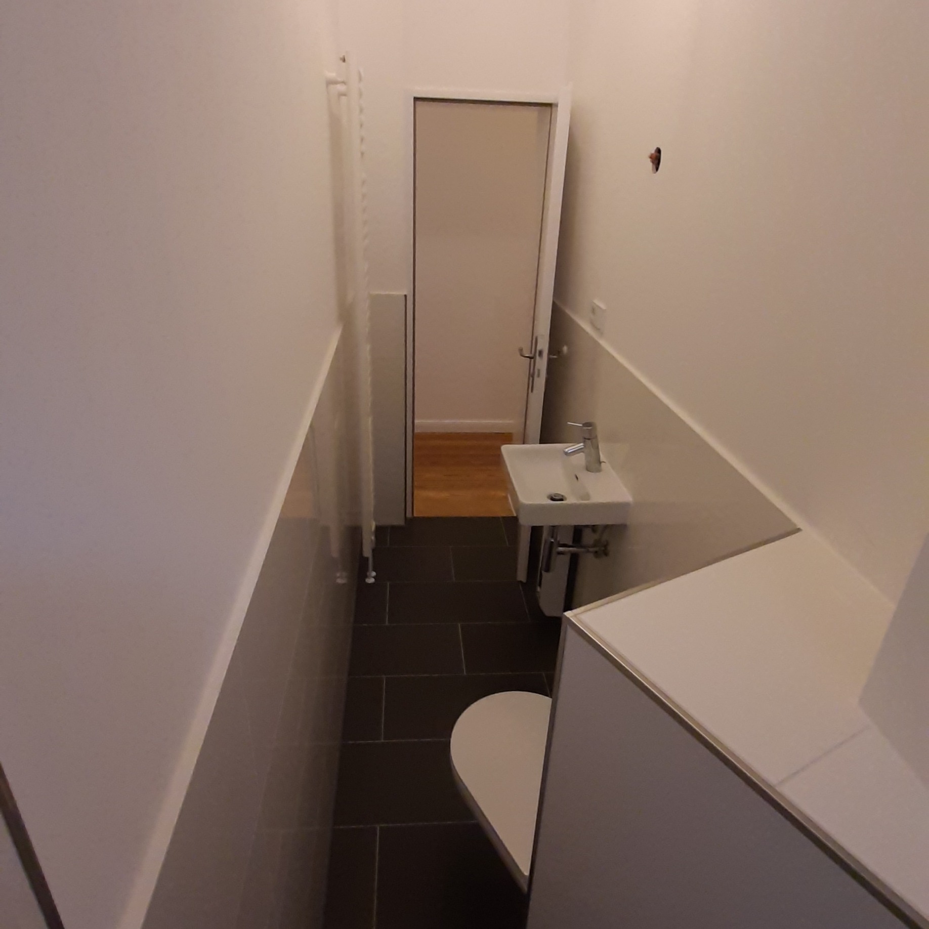 Wąska toaleta z ciemną podłogą, białą ceramiką i grzejnikiem drabinkowym, widok w głąb pomieszczenia na drewnianą podłogę w sąsiednim pokoju.