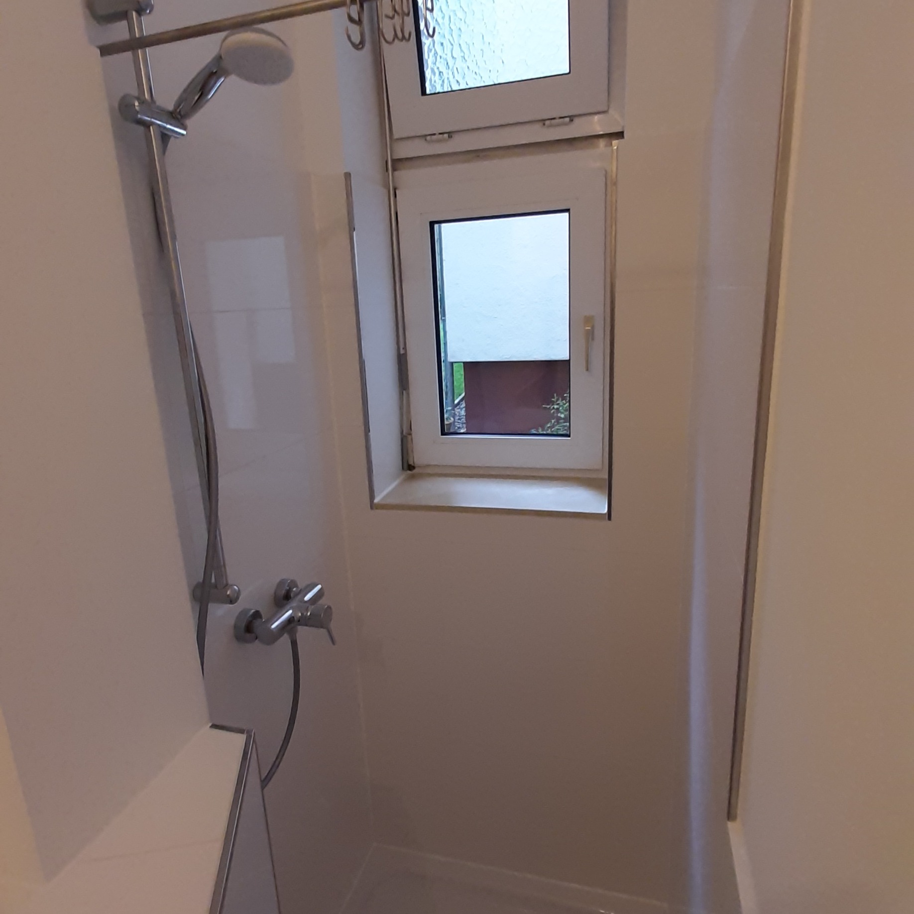 Biała łazienka z prysznicem i oknem, widok na zewnątrz, proste wykończenie.