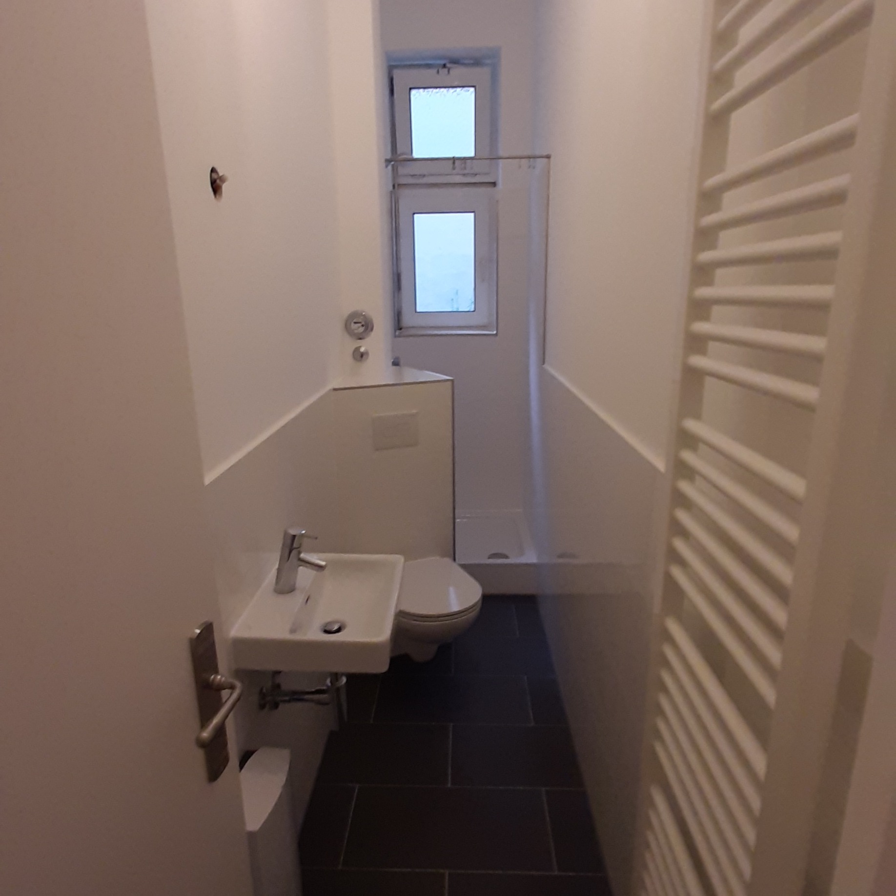 Wąska łazienka z ciemną podłogą z płytek, białą umywalką narożną, toaletą i kabiną prysznicową w tle, widoczny grzejnik drabinkowy po prawej stronie i otwór po kołku w ścianie.