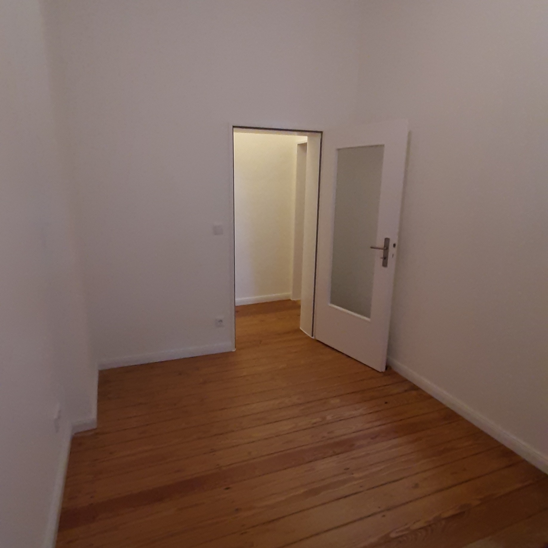 Pusty pokój z drewnianą podłogą i otwartymi białymi drzwiami z szybą, widok na kolejne pomieszczenie.