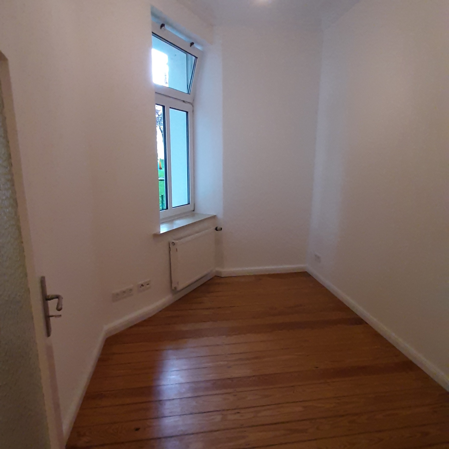 Pusty pokój z drewnianą podłogą, białymi ścianami i oknem z widokiem na zieleń, widoczne gniazdka elektryczne i grzejnik pod oknem.