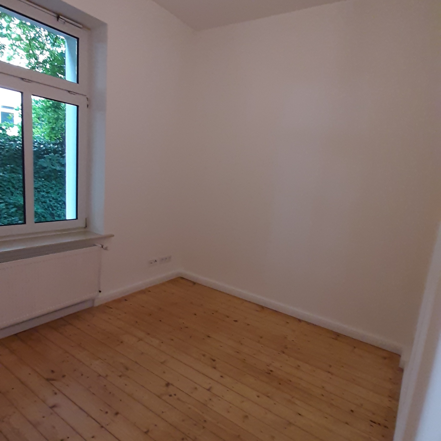 Pusty pokój z drewnianą podłogą i białymi ścianami po malowaniu, widok na okno z zielenią na zewnątrz.