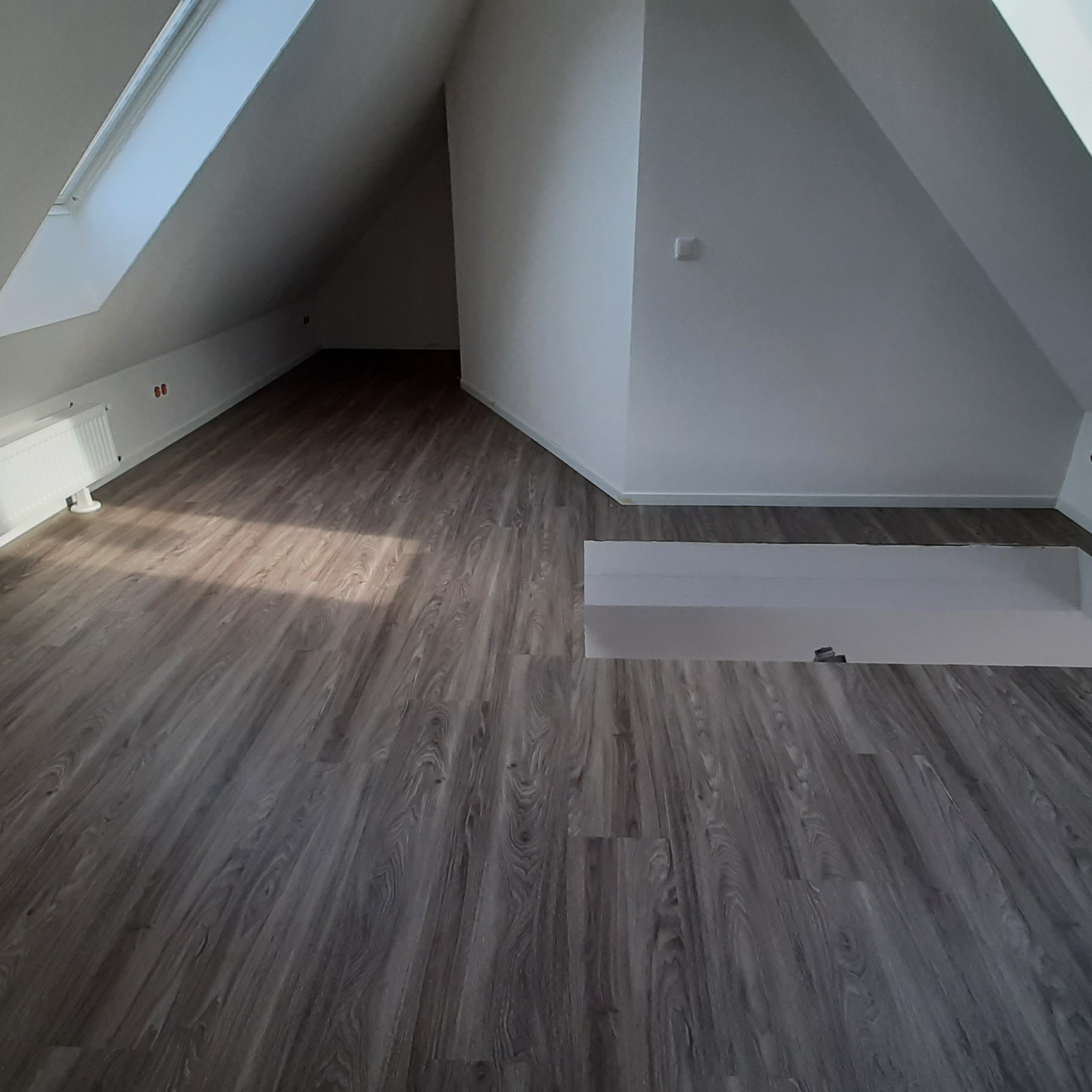 Poddasze z nowo ułożonymi panelami podłogowymi imitującymi drewno, widoczne okno dachowe i białe ściany, brak mebli.