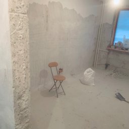 Markbud - Pokój w trakcie remontu: surowe ściany z resztkami starej farby, składane krzesło i worek gipsu. Widoczne ślady prac przygotowawczych na ścianach.