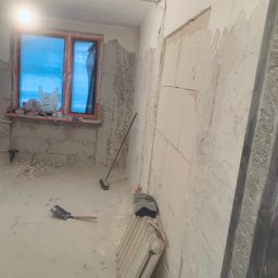 Markbud - Wnętrze w trakcie remontu: ściany z częściowo nałożoną gładzią, okno z niebieską folią, grzejnik, łopata i miotła. Widoczne ślady prac budowlanych.