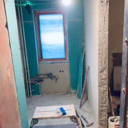 Markbud - Remont łazienki: widok na wnętrze z oknem, ściany przygotowane pod płytki, widoczne rury i instalacje. Na pierwszym planie narzędzia i rękawice robocze.