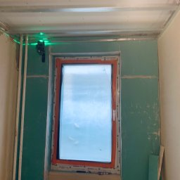 Markbud - Wnętrze w trakcie prac wykończeniowych: okno, płyty gipsowo-kartonowe na ścianach, metalowy stelaż sufitu podwieszanego i poziomica laserowa.