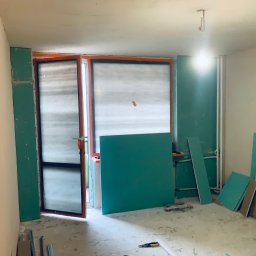 Markbud - Wnętrze w trakcie remontu: płyty gipsowe przy oknach i ścianach, surowe ściany, narzędzia na podłodze. Przygotowanie do wykończenia, widok na okno z pomarańczową ramą.