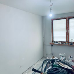 Markbud - Świeżo pomalowana ściana w pokoju z oknem i narzędziami. Widoczne worki z zaprawą i lampa wisząca na kablu. Ściana gładka, biała.