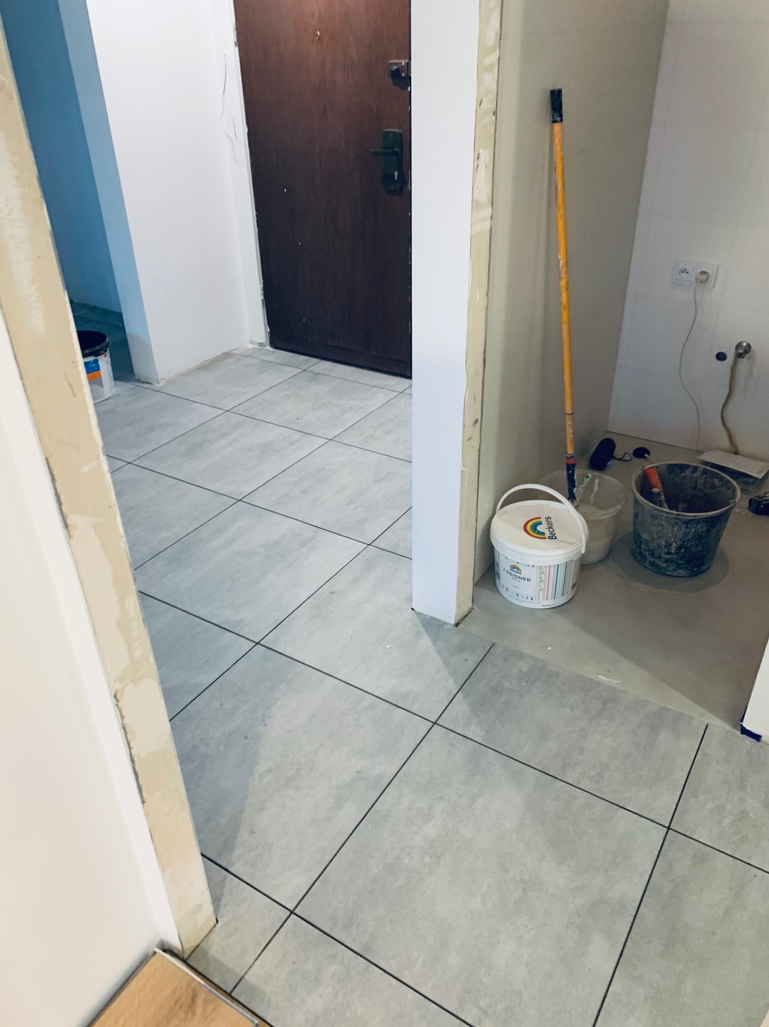 Ukończona podłoga z szarych płytek w korytarzu, widoczne narzędzia i akcesoria malarskie, drzwi wejściowe w tle. Widok z perspektywy wnętrza.