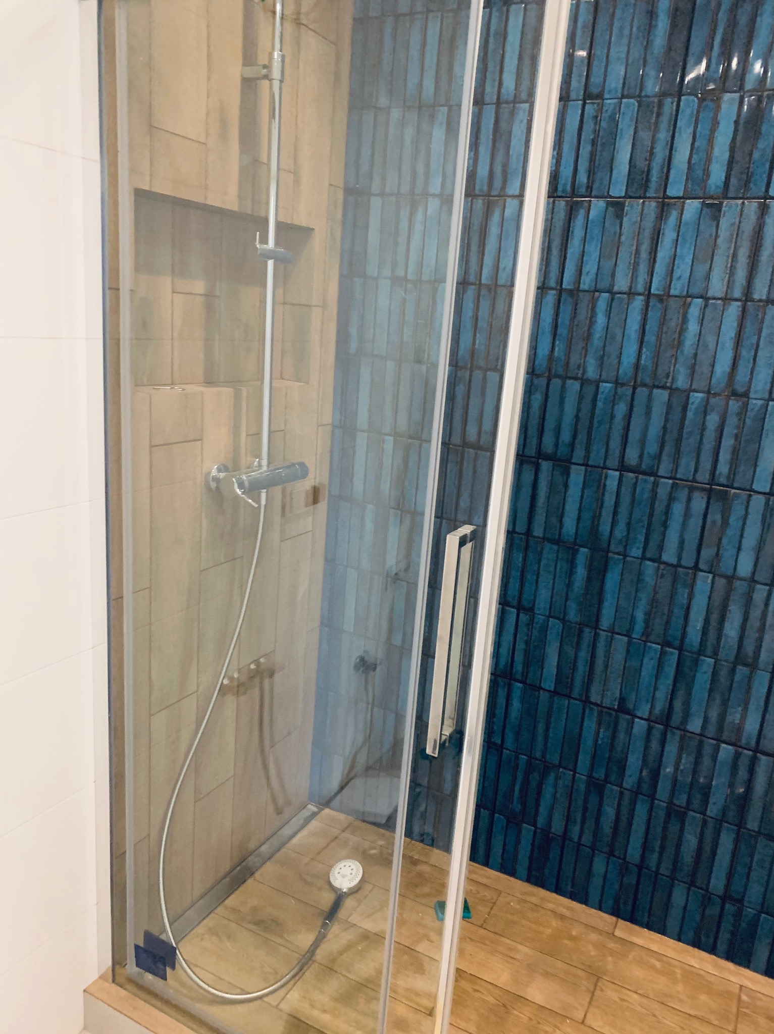 Kabina prysznicowa z niebieskimi płytkami ułożonymi pionowo, drewnopodobna podłoga i armatura. Widok z boku przez szkło.