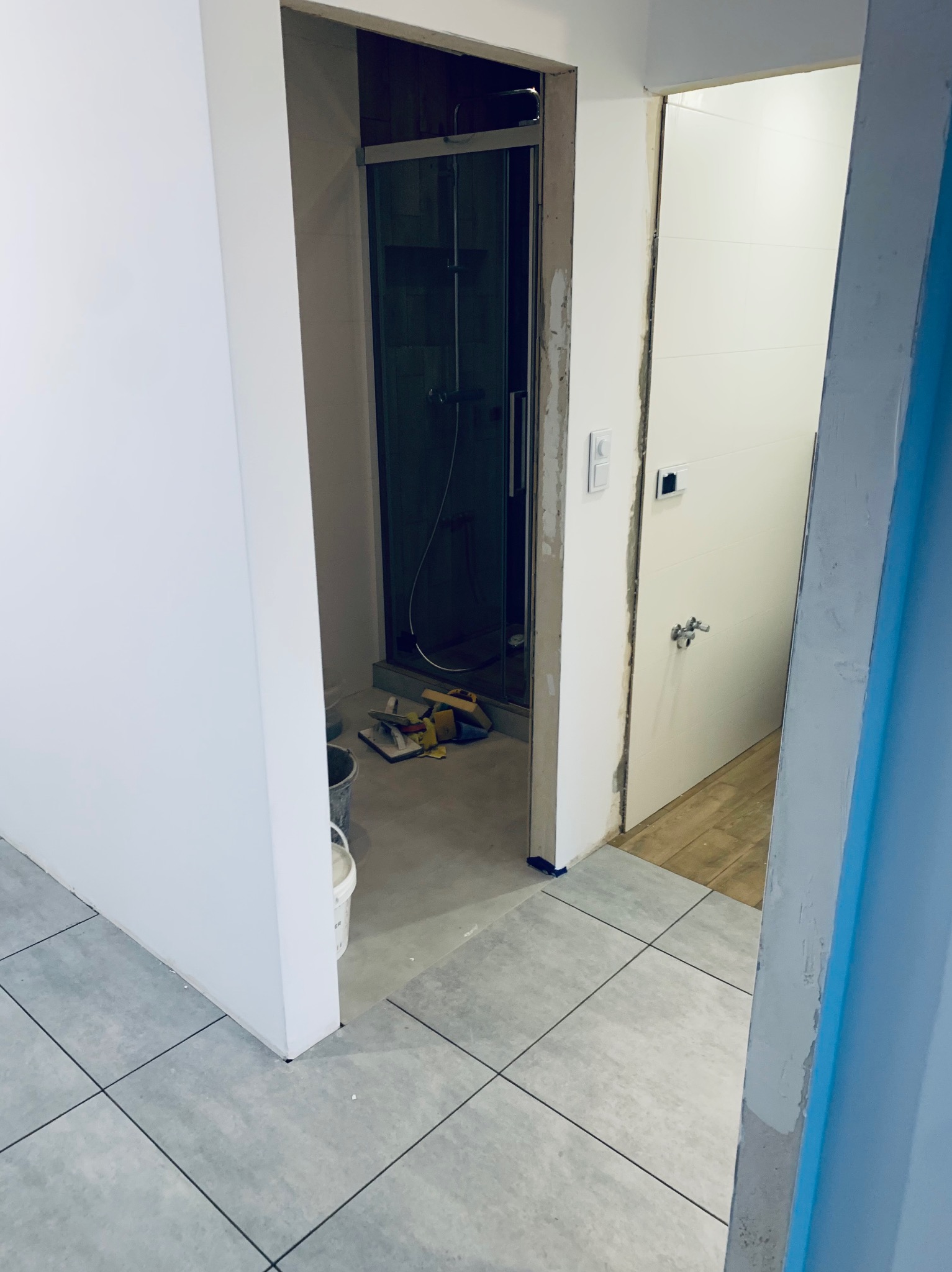 Remont łazienki w trakcie: ułożone płytki podłogowe, widoczna kabina prysznicowa z narzędziami, ściany przygotowane pod malowanie. Fragment drzwi z klamką.