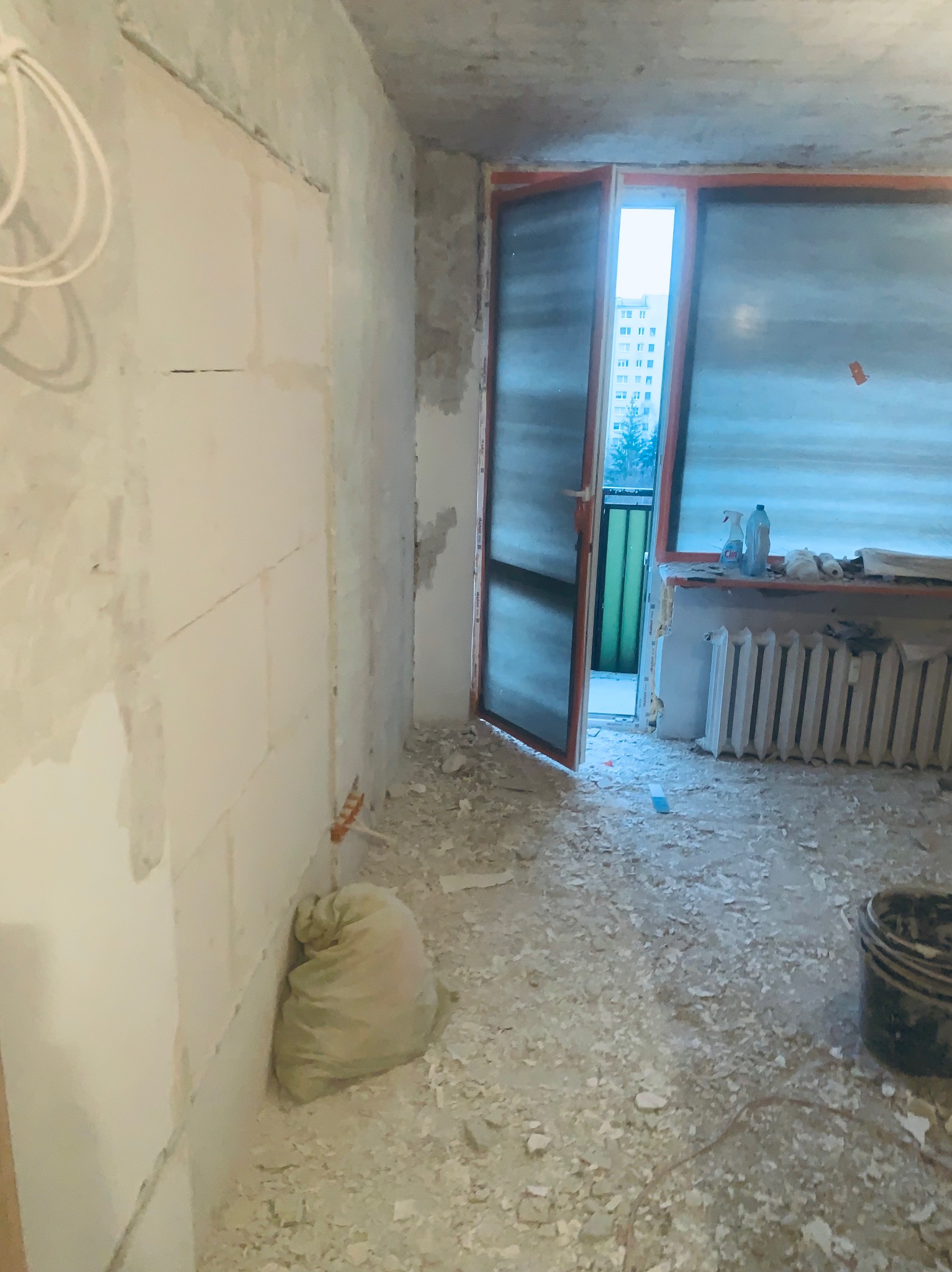 Wnętrze w trakcie remontu: surowe ściany, otwarte drzwi balkonowe, gruz na podłodze, wiadro i worek. Widoczny grzejnik i instalacja elektryczna.