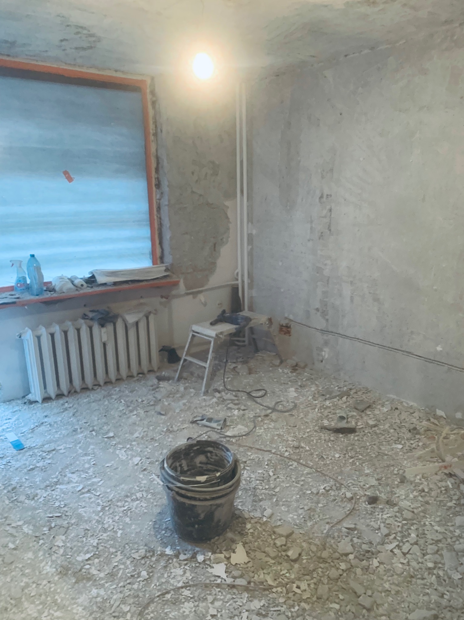 Pokój w trakcie remontu: resztki starej farby na podłodze, ściany przygotowane do gładzi, wiadro na pierwszym planie, narzędzia na stołku.