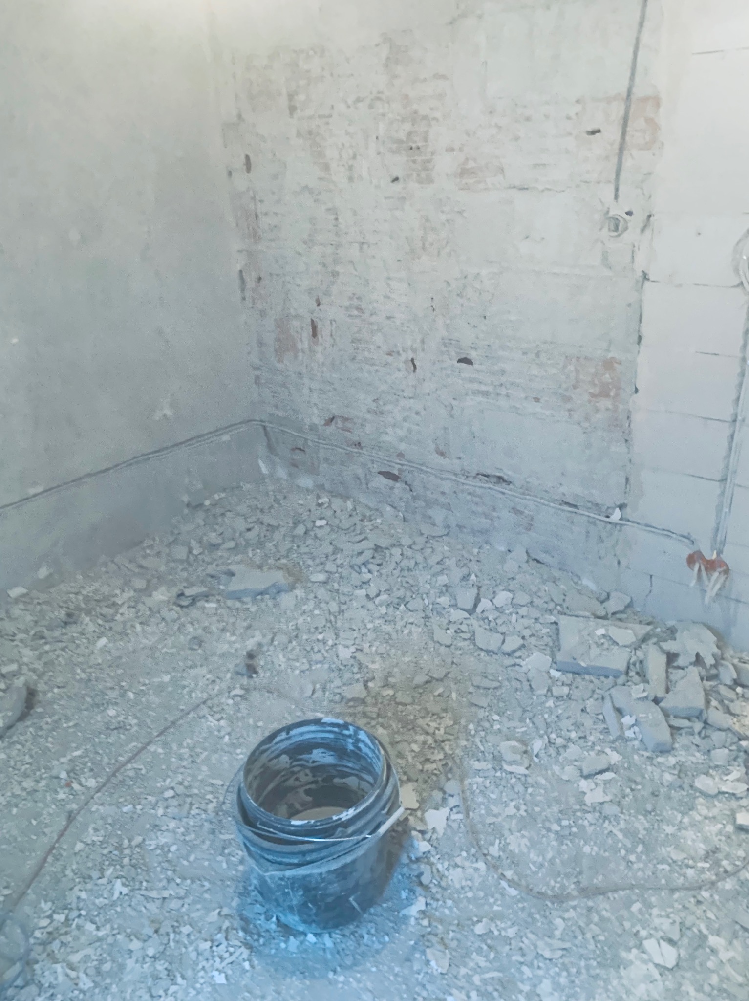 Wnętrze w trakcie remontu: fragment ściany z odsłoniętą cegłą, widoczne przewody elektryczne i gruz na podłodze. Na pierwszym planie wiadro.