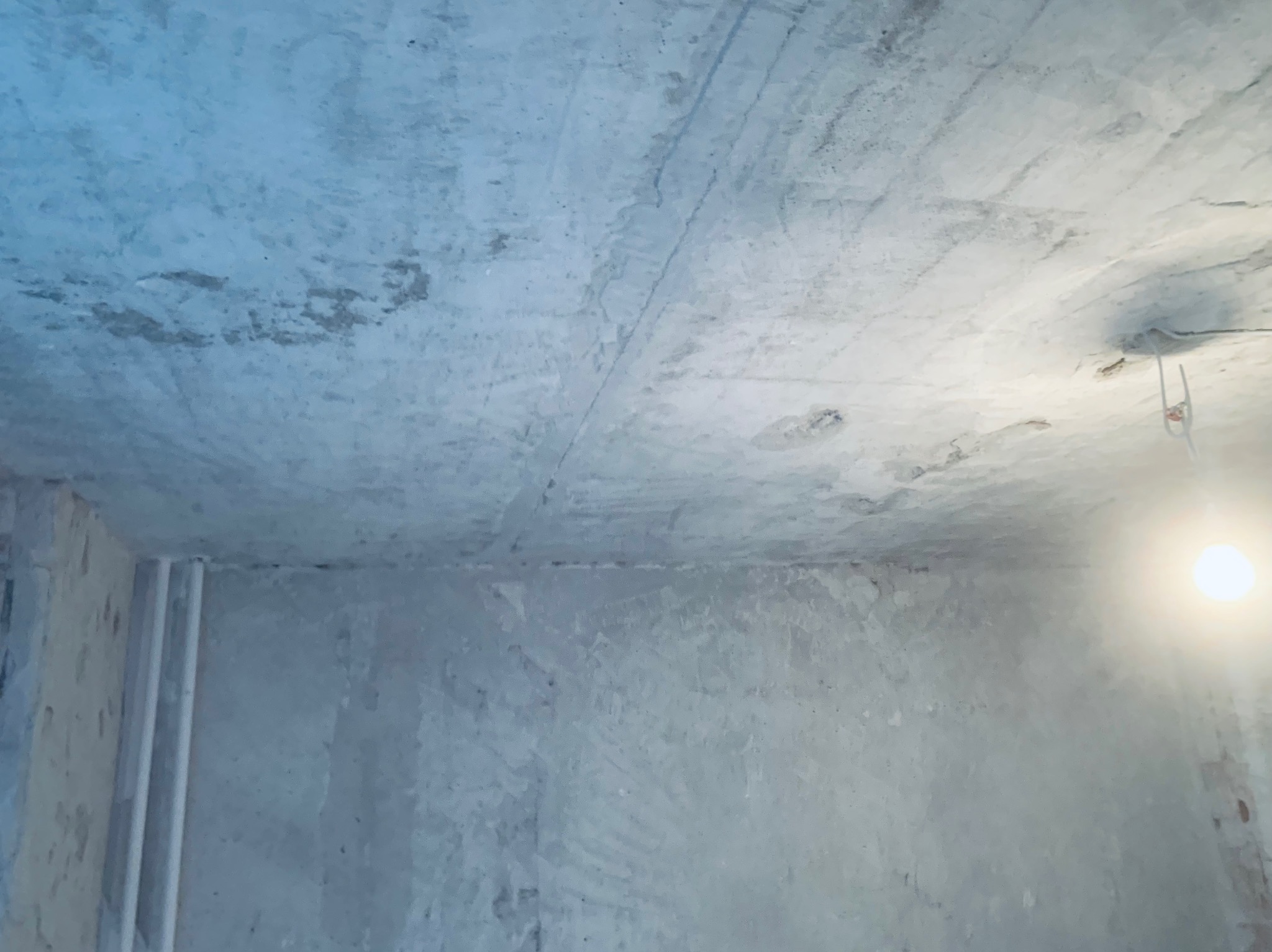 Surowe ściany i sufit po tynkowaniu w trakcie remontu, widoczne rury przy ścianie i prowizoryczne oświetlenie żarówką. Szare, nierówne powierzchnie.
