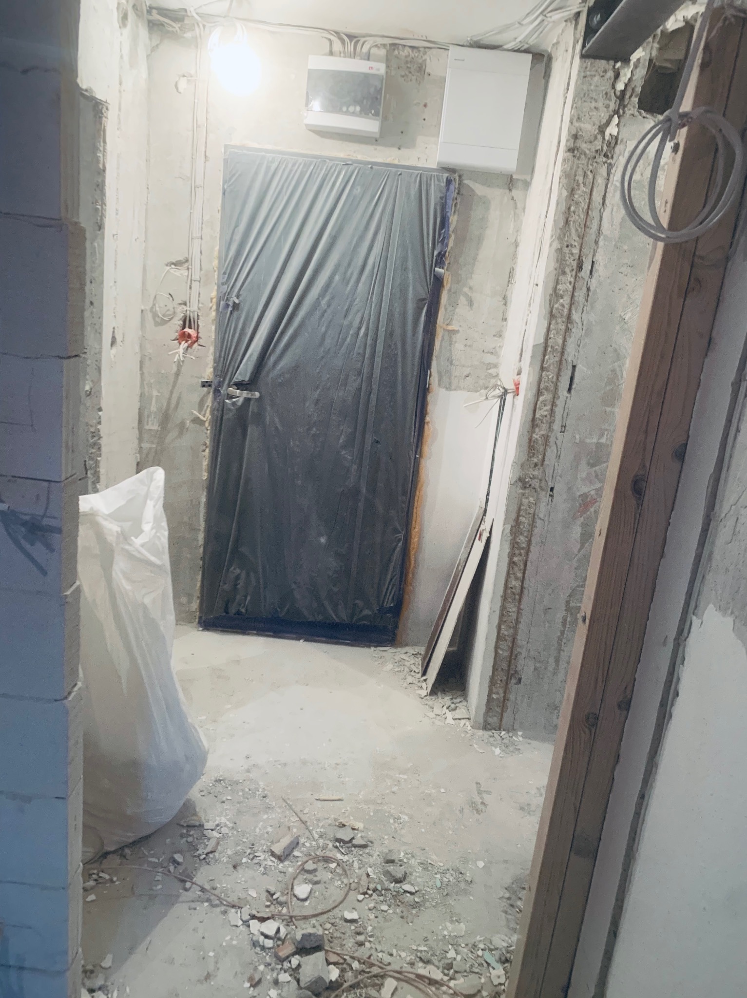 Wnętrze w trakcie remontu: zabezpieczone folią drzwi, odsłonięte przewody elektryczne, surowe ściany i gruz na podłodze. Widoczny worek z materiałem budowlanym.