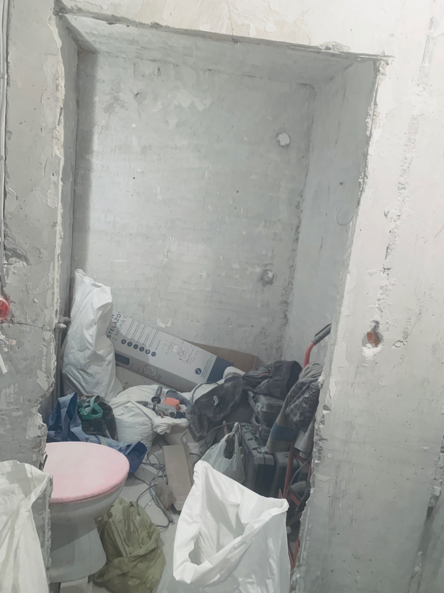 Wnęka w trakcie remontu z widocznymi surowymi ścianami, instalacjami i materiałami budowlanymi. Widoczny stelaż podtynkowy do WC i narzędzia.