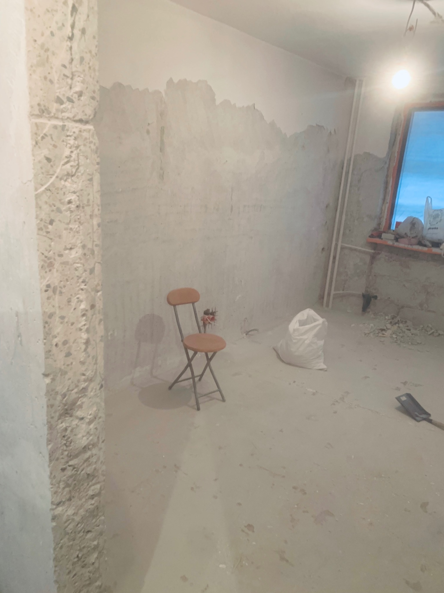 Pokój w trakcie remontu: surowe ściany z resztkami starej farby, składane krzesło i worek gipsu. Widoczne ślady prac przygotowawczych na ścianach.