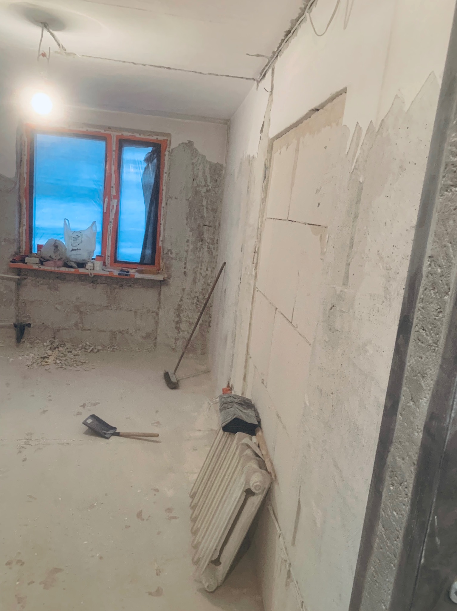 Wnętrze w trakcie remontu: ściany z częściowo nałożoną gładzią, okno z niebieską folią, grzejnik, łopata i miotła. Widoczne ślady prac budowlanych.