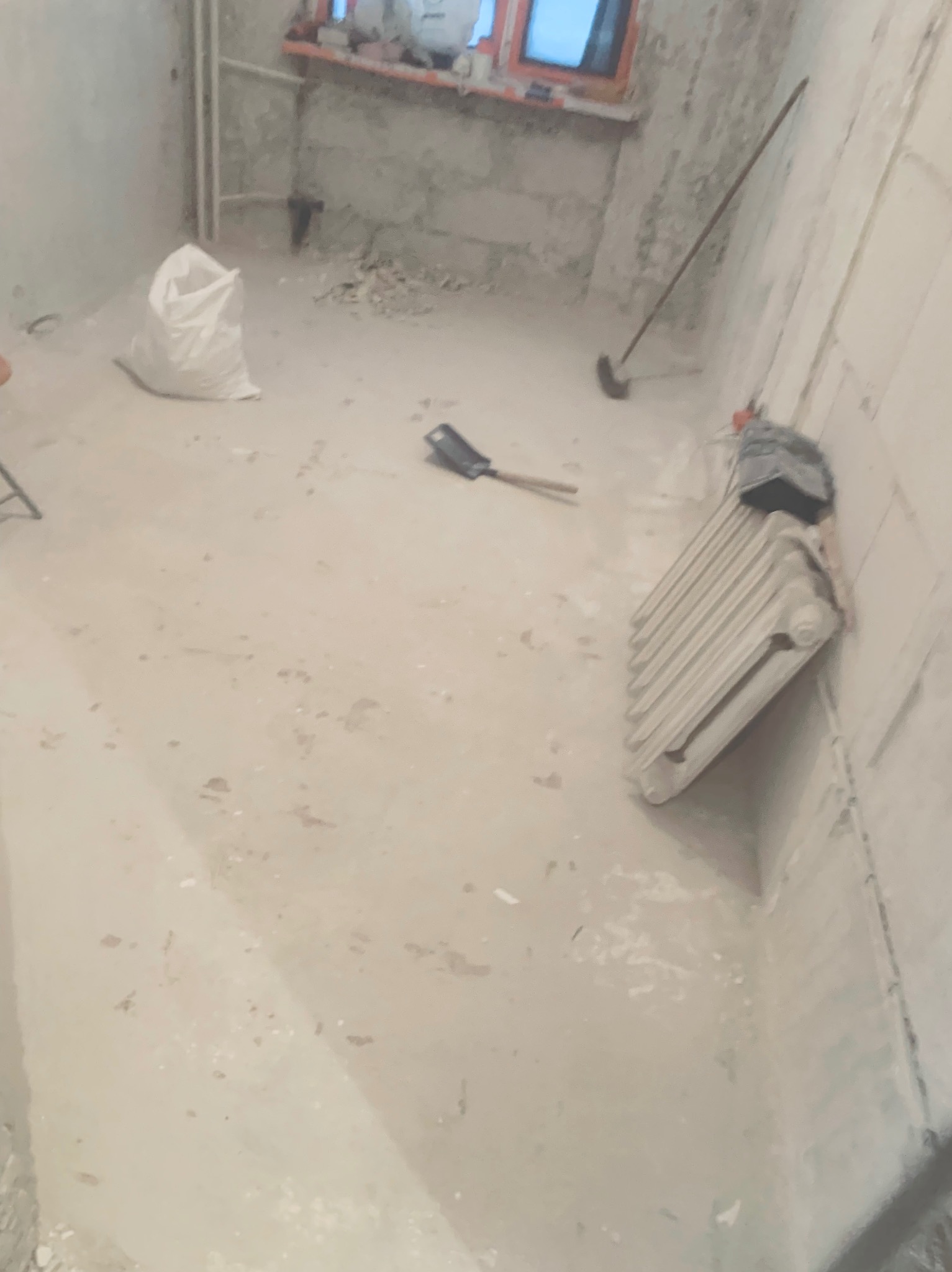 Wnętrze w trakcie remontu: surowe ściany, worek z materiałami, grzejnik przy ścianie, narzędzia. Widoczne ślady prac budowlanych.