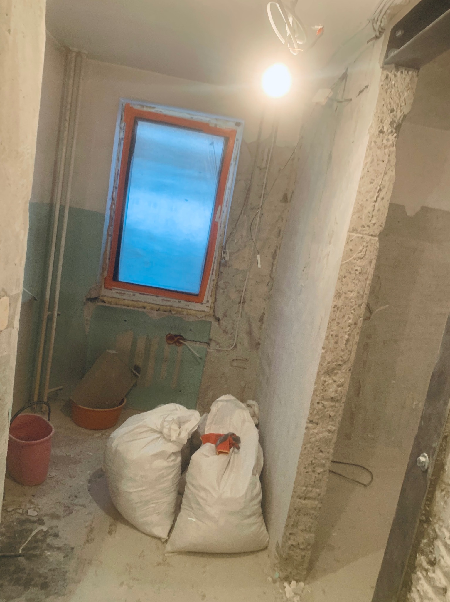 Wnętrze w trakcie remontu: gruz w workach, odkryte instalacje elektryczne przy oknie z pomarańczową ramą, ściany w stanie surowym, widoczne rury i pozostałości po tynku.