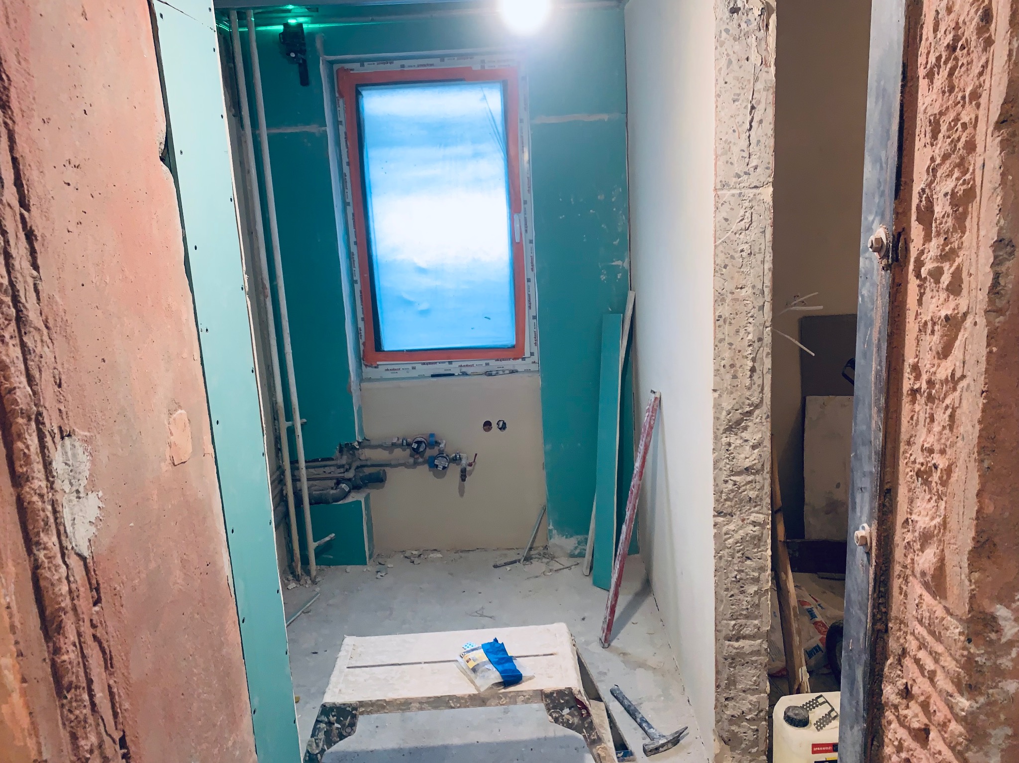 Remont łazienki: widok na wnętrze z oknem, ściany przygotowane pod płytki, widoczne rury i instalacje. Na pierwszym planie narzędzia i rękawice robocze.