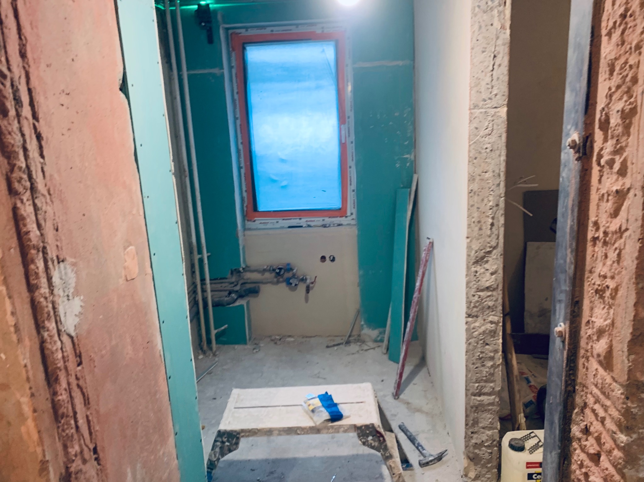 Wnętrze łazienki w trakcie remontu: ściany z płyt gipsowo-kartonowych, okno z pomarańczową ramą, widoczne rury instalacji wodnej i narzędzia na podwyższeniu.