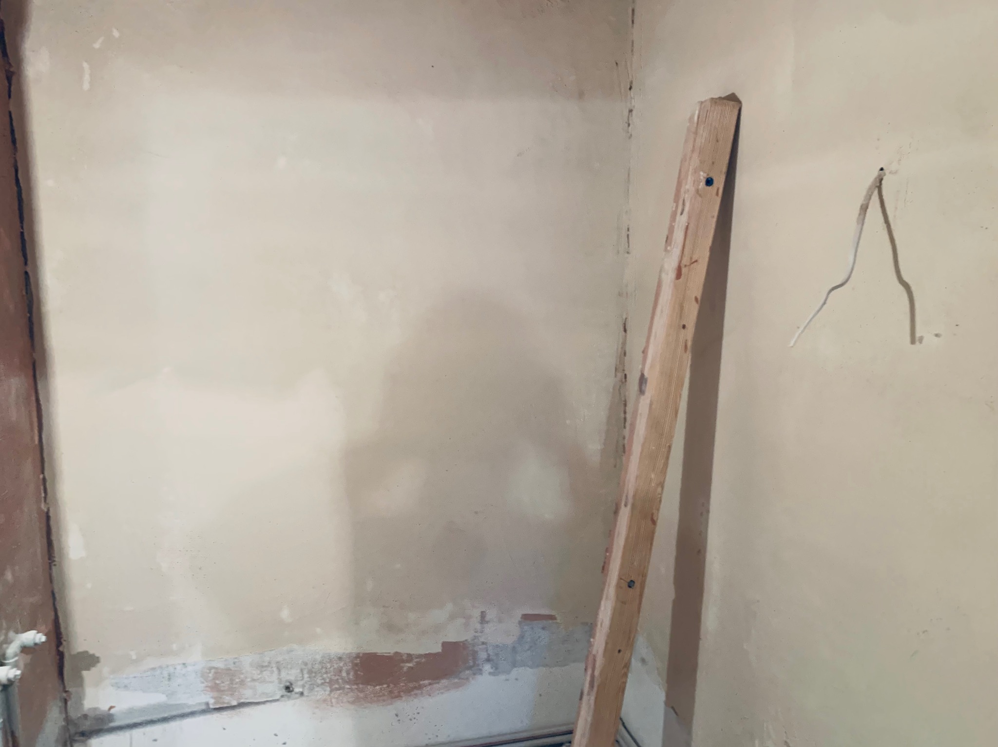 Fragment wnętrza w trakcie prac wykończeniowych: ściana po nałożeniu gładzi, widoczne niedoskonałości i drewniana listwa oparta o ścianę. W tle rury.