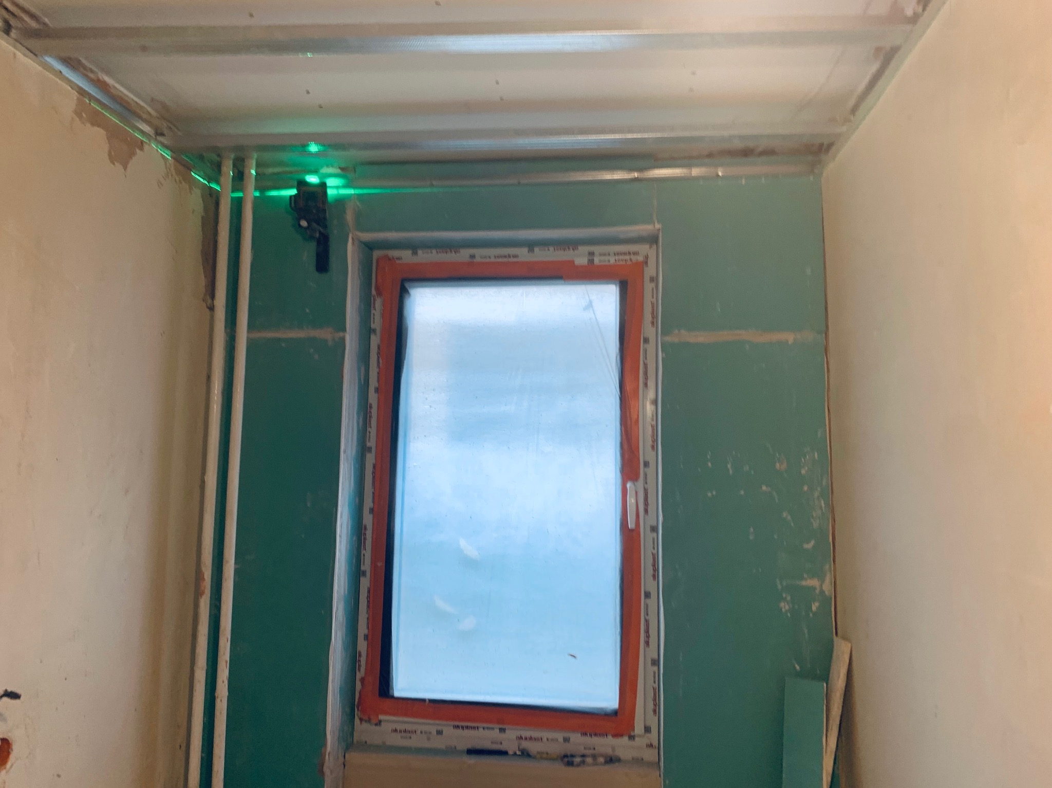 Wnętrze w trakcie prac wykończeniowych: okno, płyty gipsowo-kartonowe na ścianach, metalowy stelaż sufitu podwieszanego i poziomica laserowa.