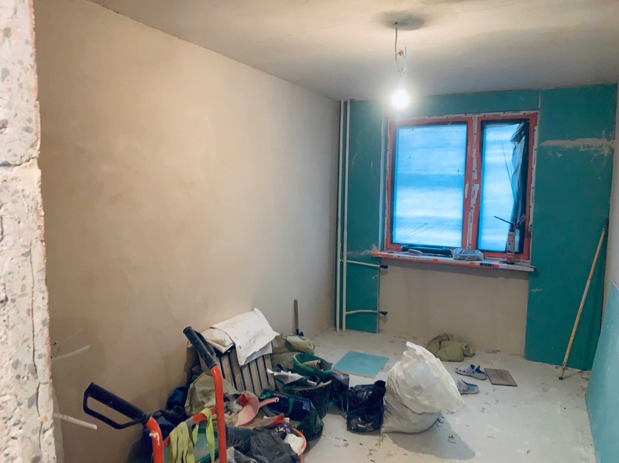 Pokój w trakcie remontu: ściana z nałożoną gładzią, okno zabezpieczone folią, płyty gipsowe na ścianie, narzędzia i worki w nieładzie. Widok na wnętrze w trakcie prac.