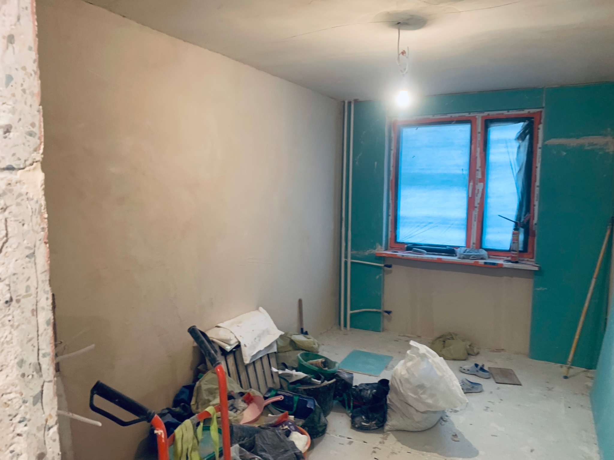 Pokój w trakcie prac wykończeniowych: ściany częściowo otynkowane, okno zabezpieczone folią, widoczne narzędzia i materiały budowlane. Fragmentarycznie położona płyta karton gips.