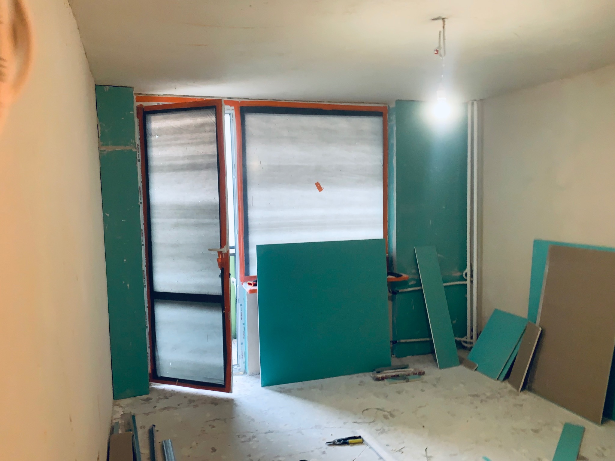Wnętrze w trakcie remontu: płyty gipsowe przy oknach i ścianach, surowe ściany, narzędzia na podłodze. Przygotowanie do wykończenia, widok na okno z pomarańczową ramą.