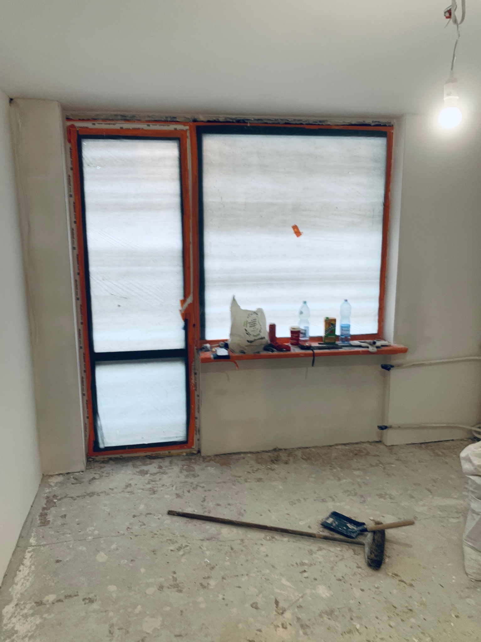 Wnętrze w trakcie prac wykończeniowych: okno zabezpieczone folią, narzędzia na podłodze, ściany przygotowane do malowania, widoczna żarówka.