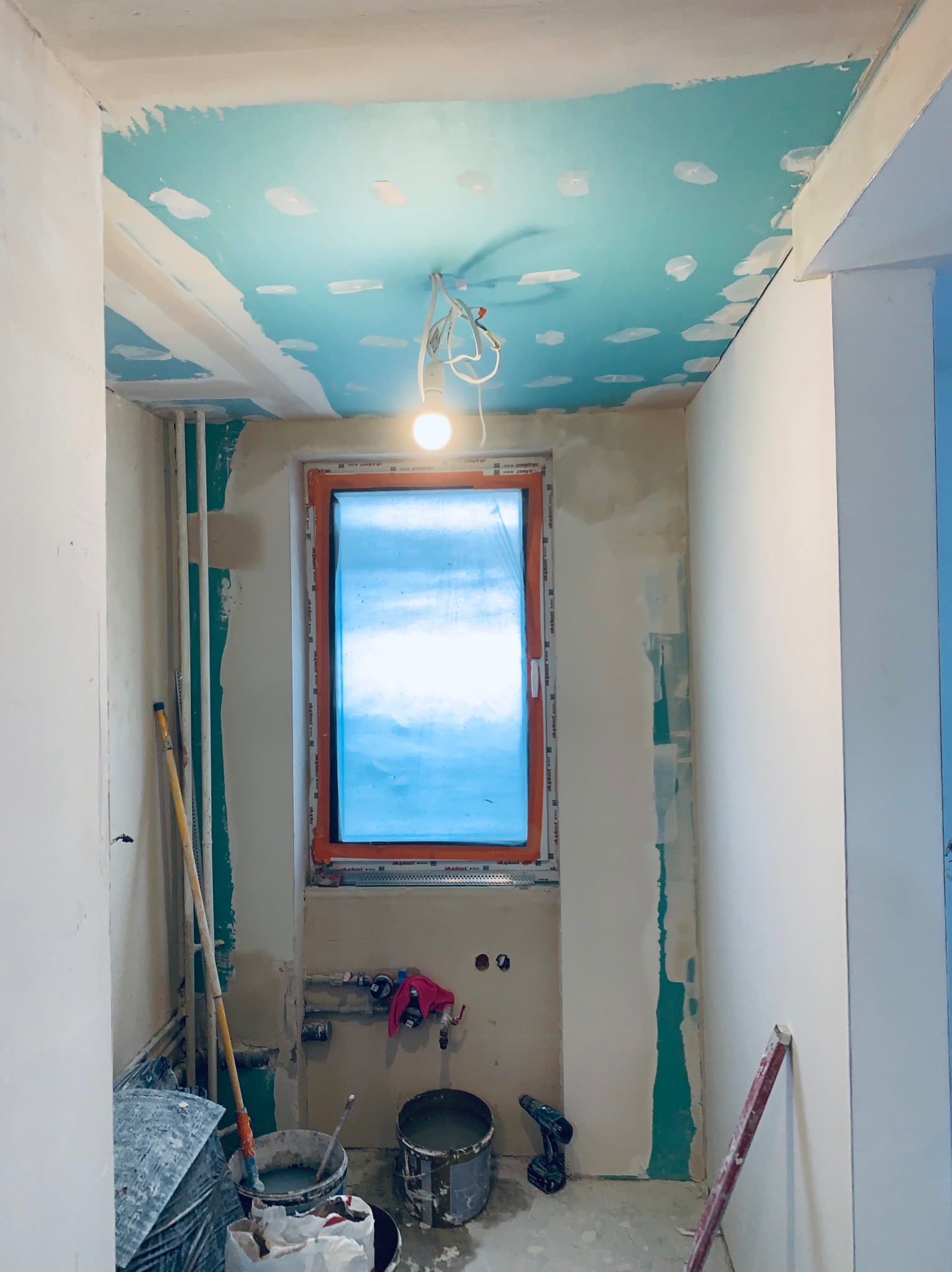 Wnętrze łazienki w trakcie remontu: surowy sufit z niebieskimi fragmentami, okno z pomarańczową ramą, ściany przygotowane pod gładź, widoczne narzędzia i wiaderka.