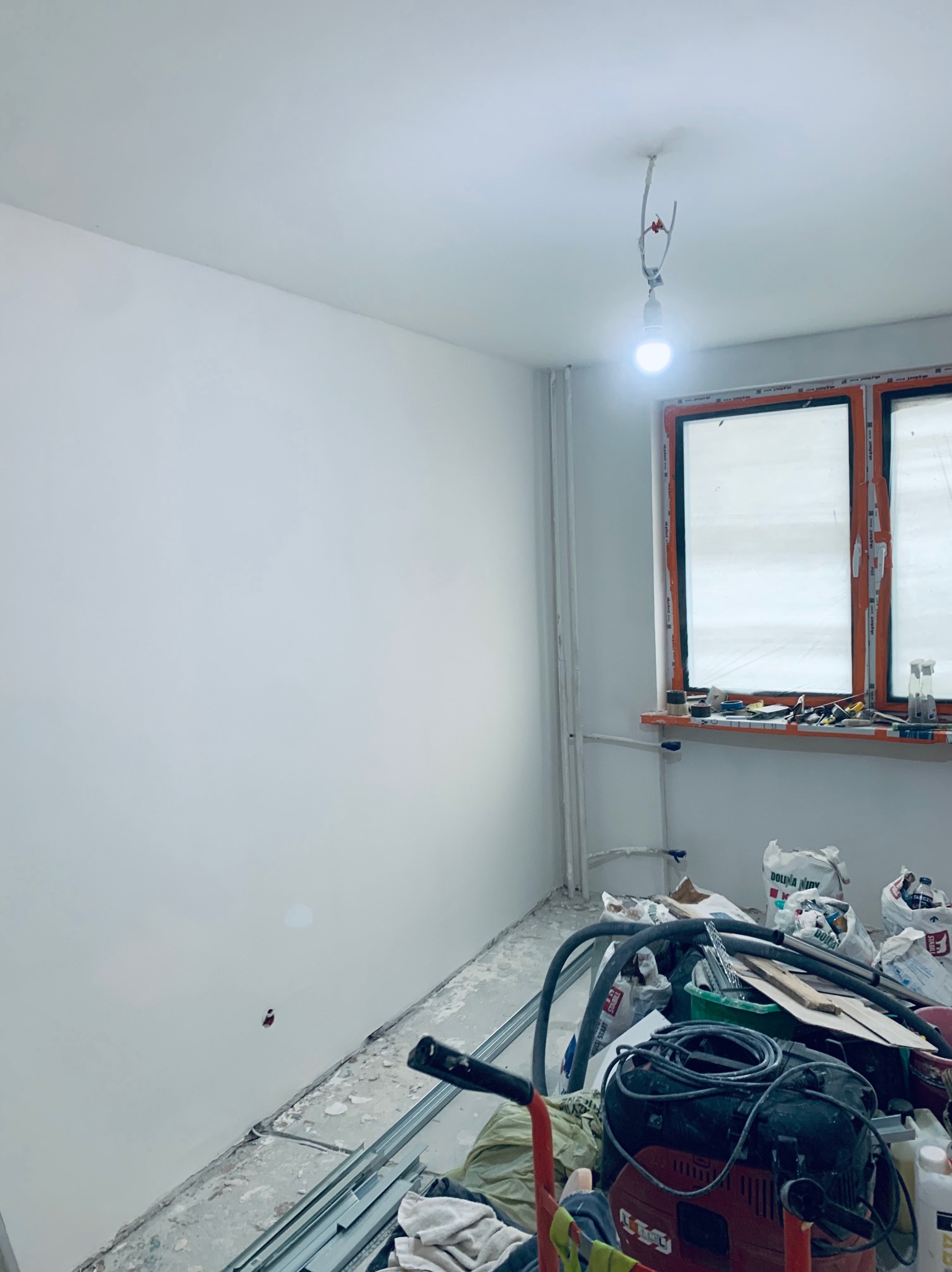 Świeżo pomalowana ściana w pokoju z oknem i narzędziami. Widoczne worki z zaprawą i lampa wisząca na kablu. Ściana gładka, biała.