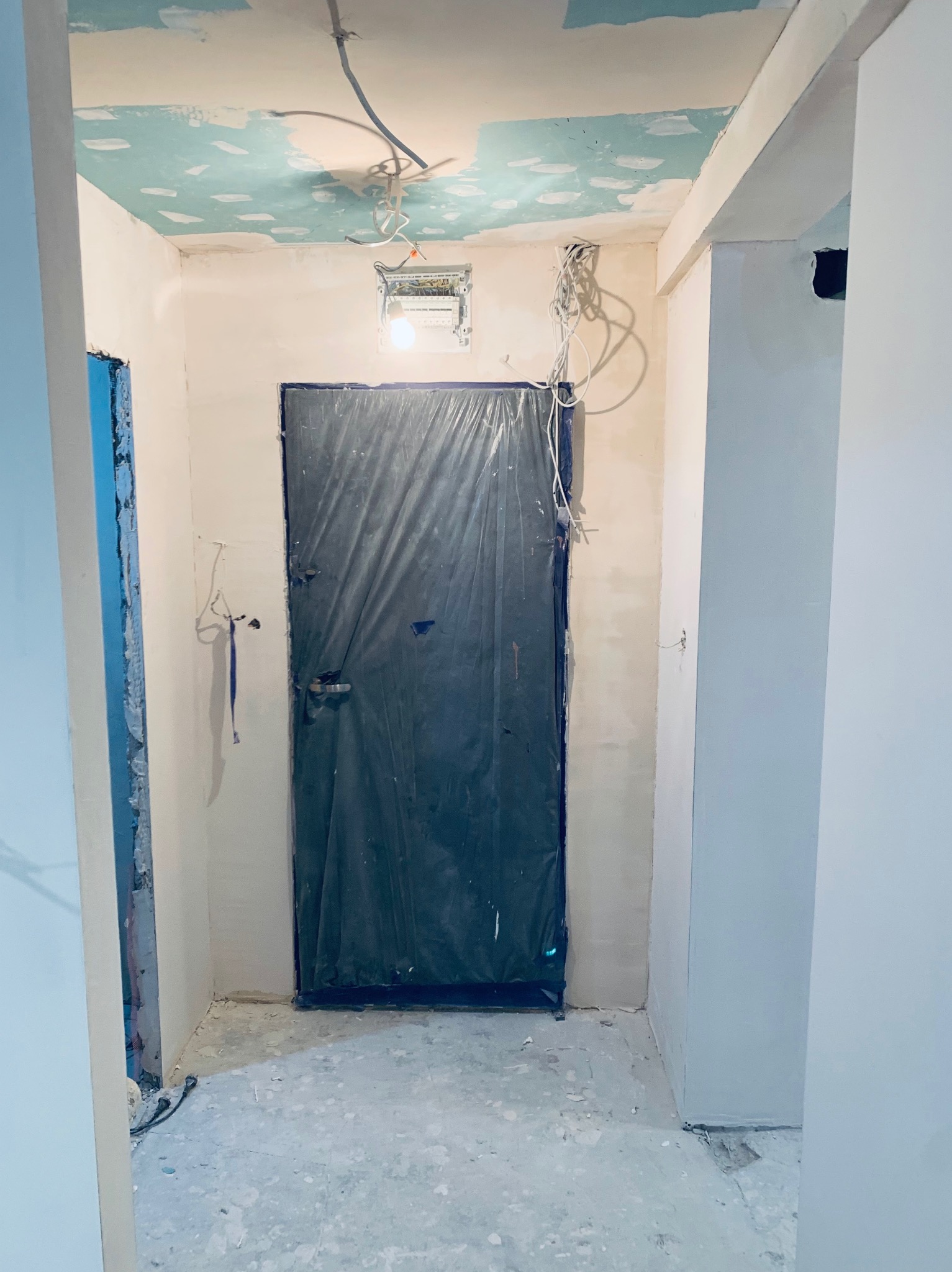 Korytarz w trakcie remontu: ściany po gładzi, sufit z fragmentami pomalowanej farby, zabezpieczone drzwi folią, widoczne przewody elektryczne.