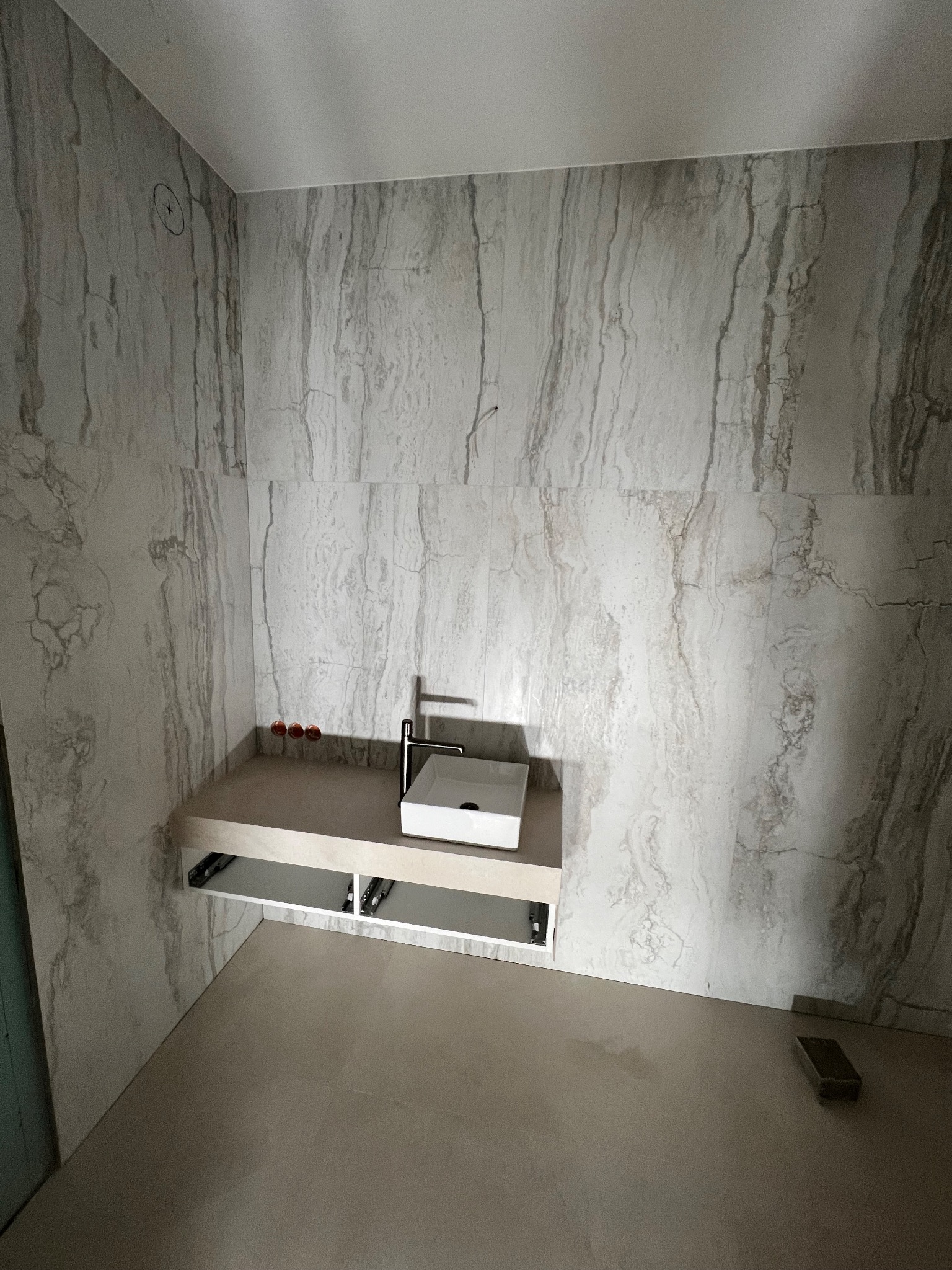 Łazienka w trakcie wykańczania, widoczne marmurowe płytki na ścianach, minimalistyczna umywalka nablatowa z chromowaną baterią, przygotowane przyłącza wodne i fragment podłogi z pojedynczą płytką.