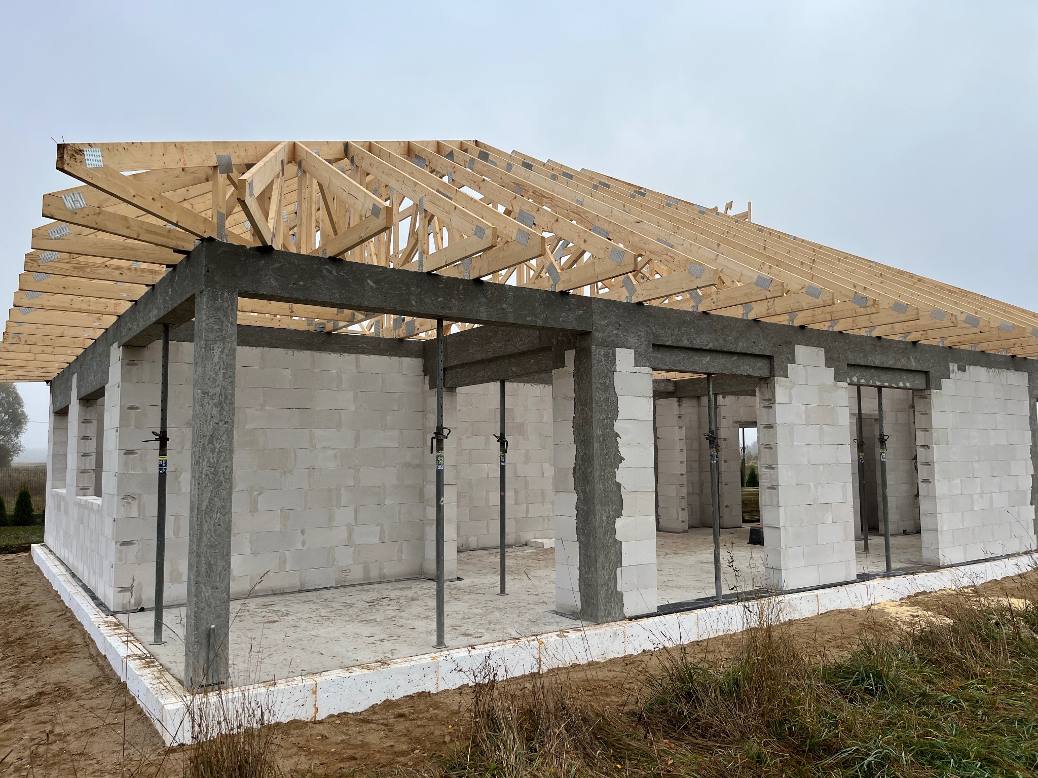 Wznoszenie surowej konstrukcji domu z betonu komórkowego, widoczne ściany, betonowe słupy wzmacniające i drewniana więźba dachowa z metalowymi łącznikami, na zewnątrz ocieplenie fundamentów.