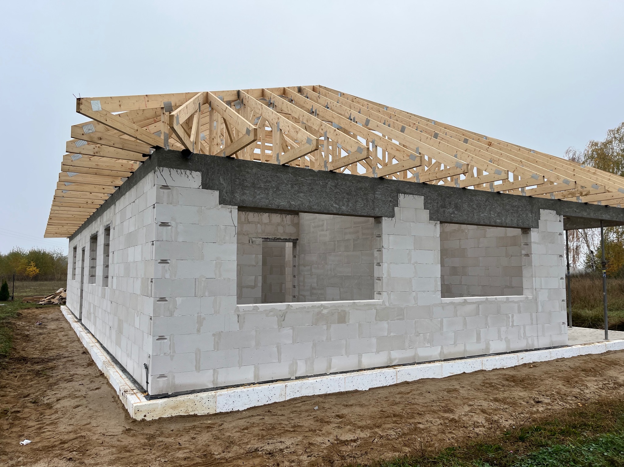 Surowy stan domu w trakcie budowy: ściany z betonu komórkowego, drewniana konstrukcja dachu z metalowymi łącznikami, fundament z izolacją termiczną, widok w pochmurny dzień.